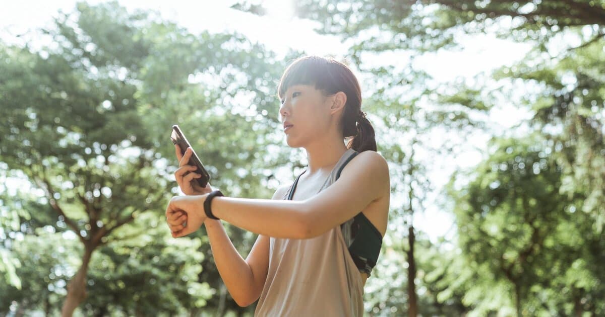 A woman in activewear holds up a smartphone while wearing a smartwatch in a sunny, tree-lined park