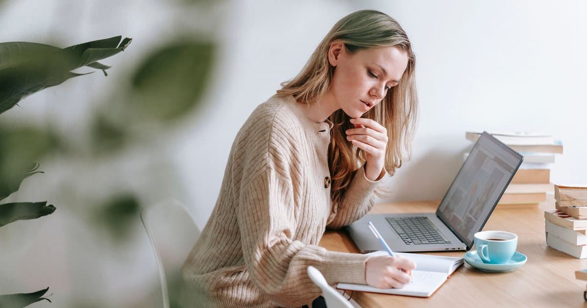 A woman in a beige sweater sits at a wooden desk with her hand on her chin, writing in a notebook next to an open laptop, a blue coffee cup, and a stack of books