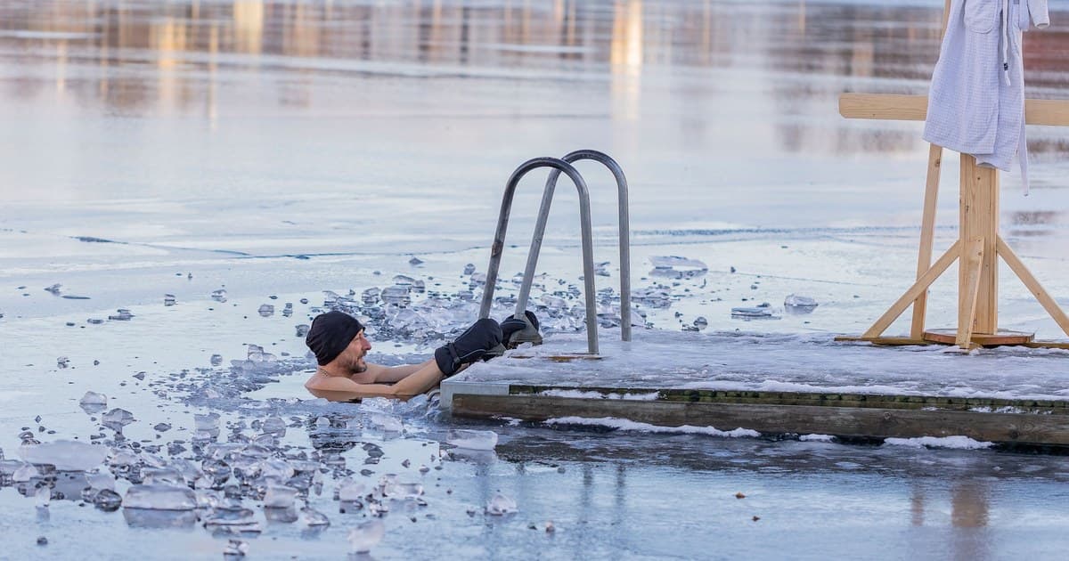 A man in a black knit hat soaks up to his shoulders in a hole cut in a frozen lake, next to a wooden dock with a metal ladder and a towel hanging on a wooden stand