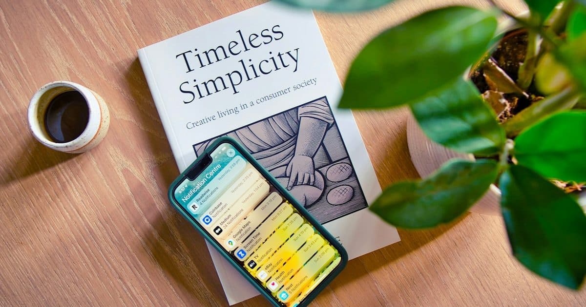 A smartphone showing notifications resting on a book titled 'Timeless Simplicity' on a wooden table, with a cup of coffee and a potted plant