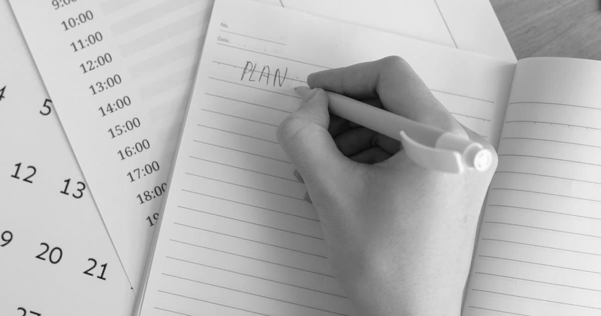 A hand writing the word PLAN in a notebook with a calendar and hourly schedule in the background