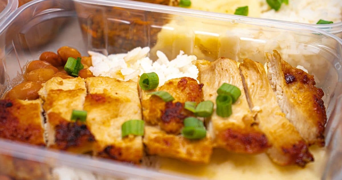 Close-up of sliced grilled chicken with white rice, beans, and chopped green onions in a clear meal-prep container