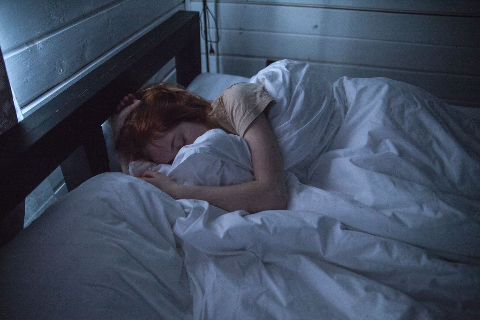 A woman with red hair sleeping peacefully under white bedding in a dim, calm bedroom