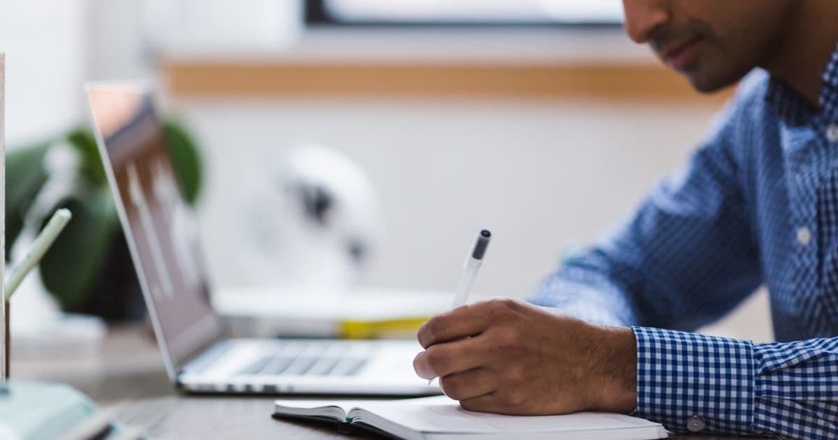 A man sits at an office desk writing in a notebook beside an open laptop, focused on a single task