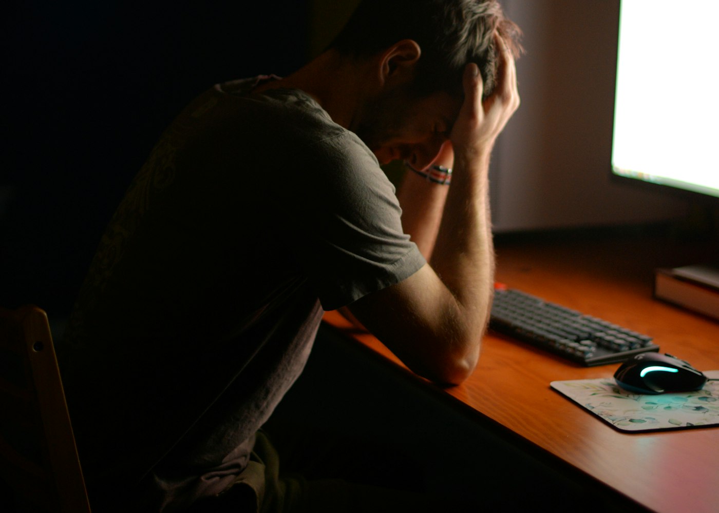 A tired man at his desk late at night with his head in his hands, lit by a computer monitor