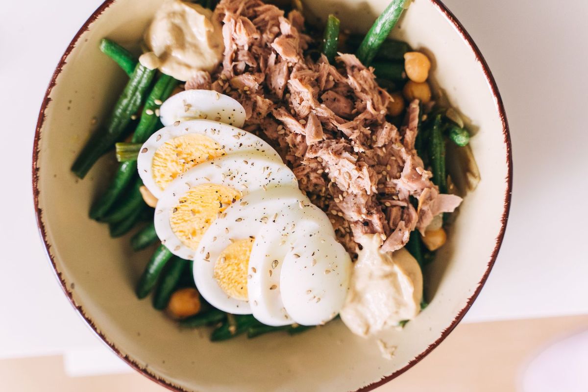 A ceramic bowl of flaked tuna, sliced hard-boiled eggs, green beans, chickpeas, and hummus on a white table
