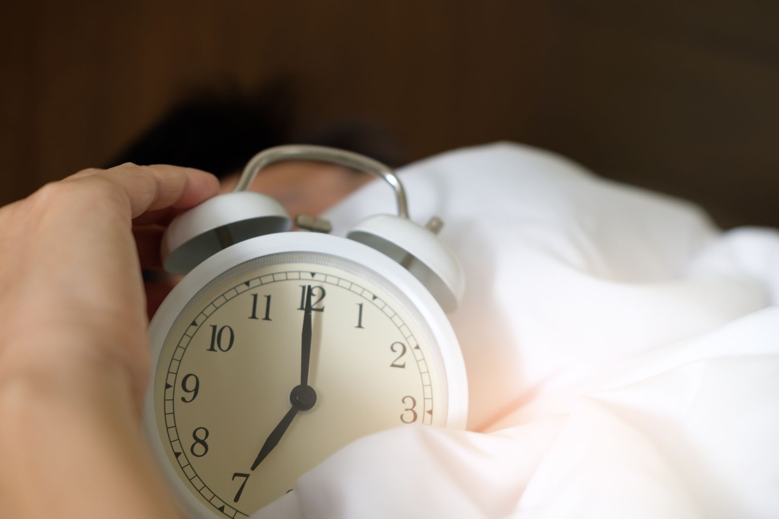 A hand reaching to silence a white twin-bell alarm clock showing 7:00 on rumpled white bedding