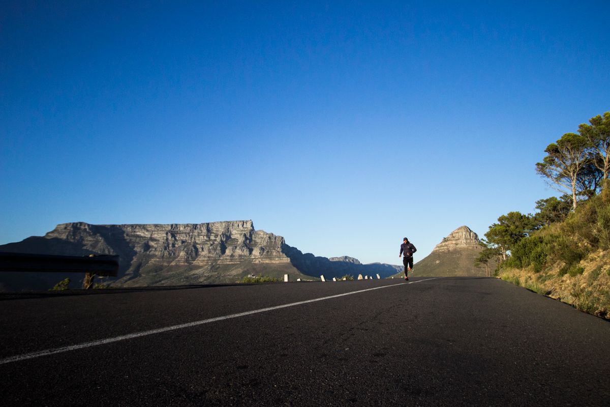 A runner in dark athletic clothing jogging on a mountain road under a clear blue sky with Table Mountain in the background